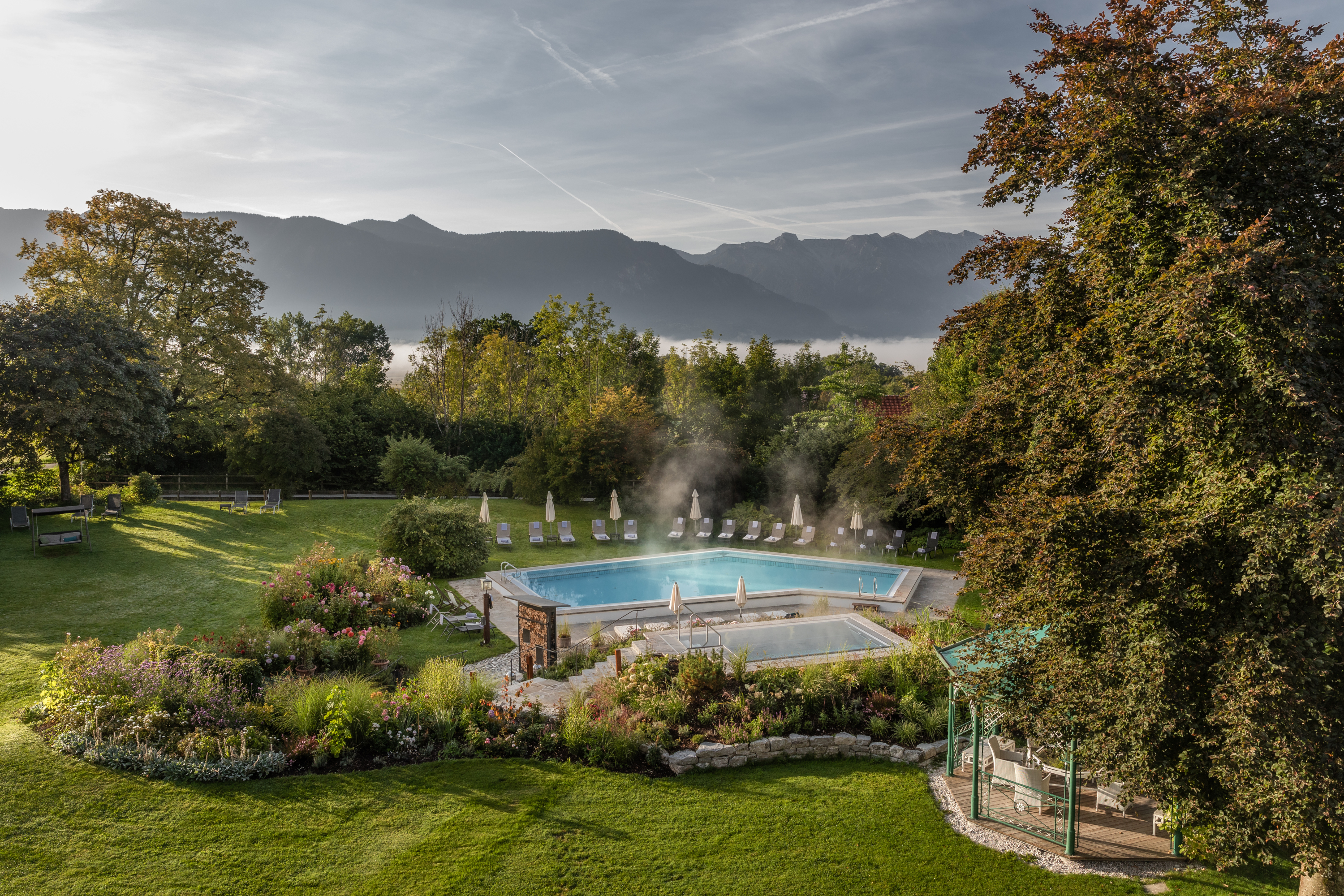 Beheizter Außenpool mit Liegestühlen im Garten eines Hotels, umgeben von blühenden Beeten, Bäumen und Bergpanorama im Hintergrund